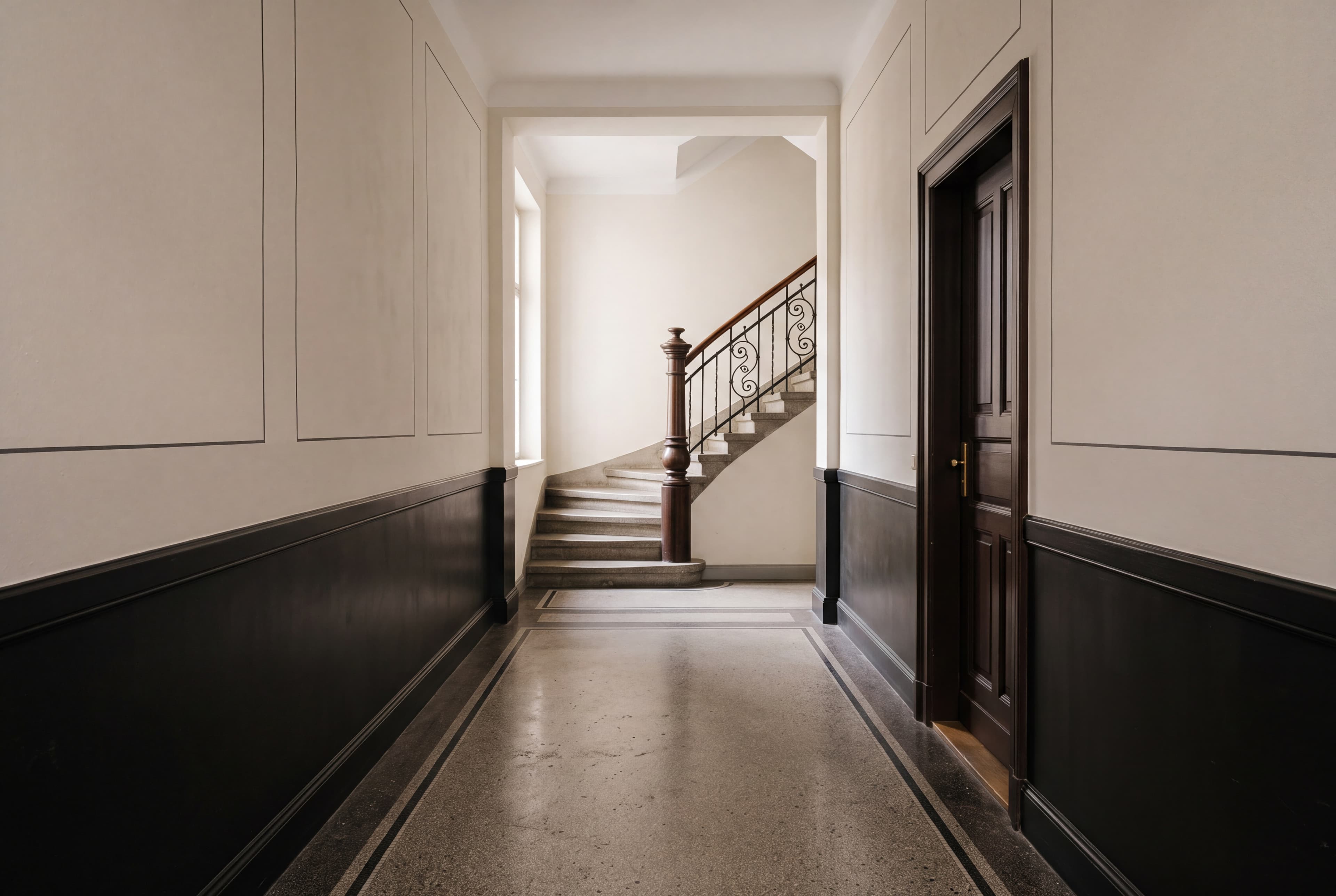 Leerer Eingangsflur eines Münchner Altbaus: Terrazzoboden mit dunkler Zierbordüre, dunkler Ölsockel unter hellen Wandfeldern, am Ende der Treppenantritt mit gedrechseltem Pfosten und schmiedeeisernem Geländer