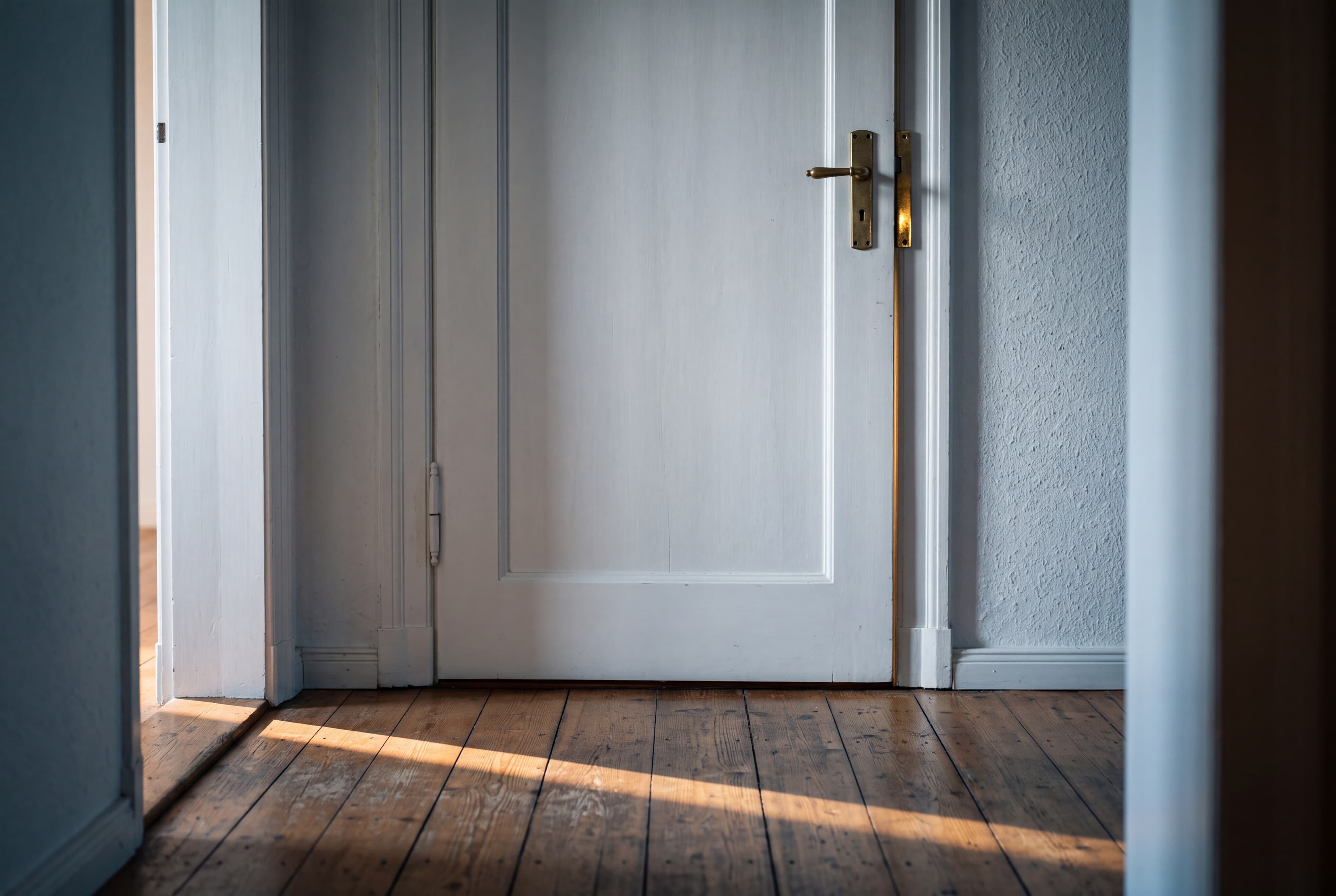 Geschlossene weiße Wohnungstür mit Messingdrücker, davor ein schmaler Lichtstreifen auf Dielen